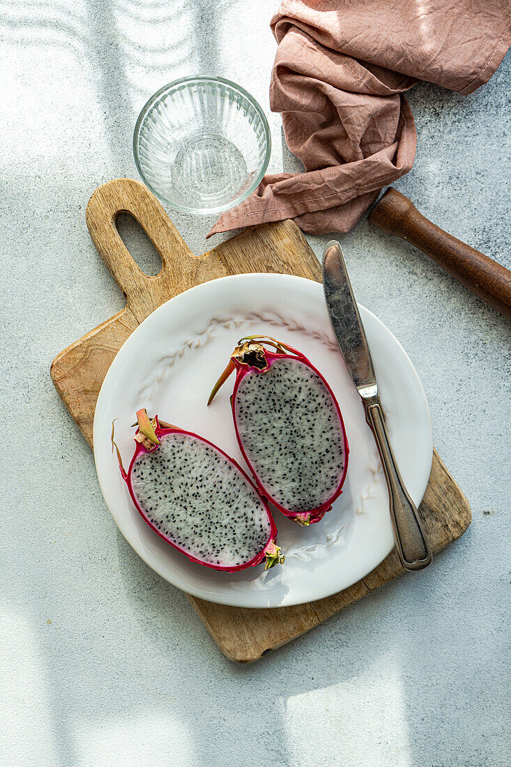 A vibrant dragonfruit sliced in half, presented on a white plate with a rustic wooden board and kitchen utensils.
