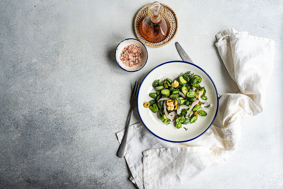 Top view of cucamelon salad with red onion, olives and spicy cheese served on white plate with fork and knife near napkin and oil on gray table in daylight