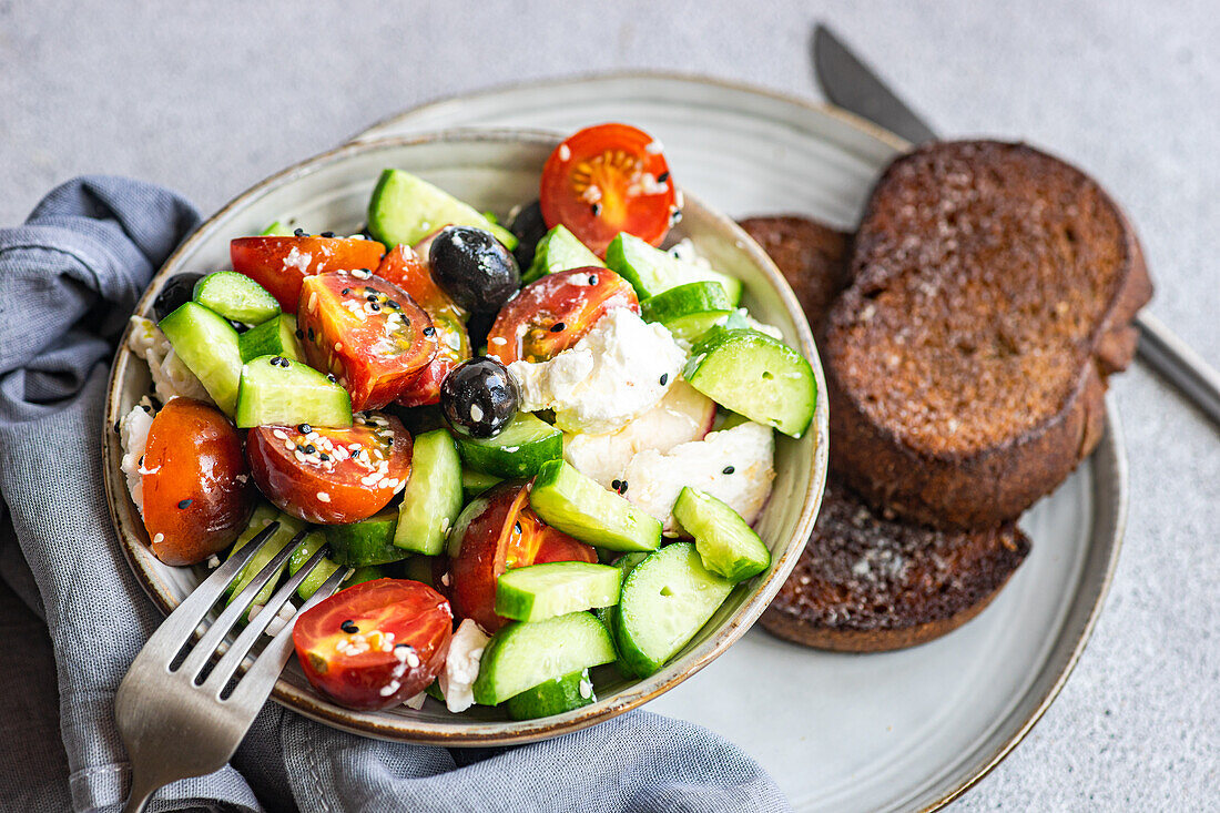 Hoher Blickwinkel auf eine Keramikschale mit leckerem, gesundem Salat, Gemüse und Brot, die mit Gurken- und Tomatenscheiben, Besteck und Stoff auf einem grauen Tisch steht