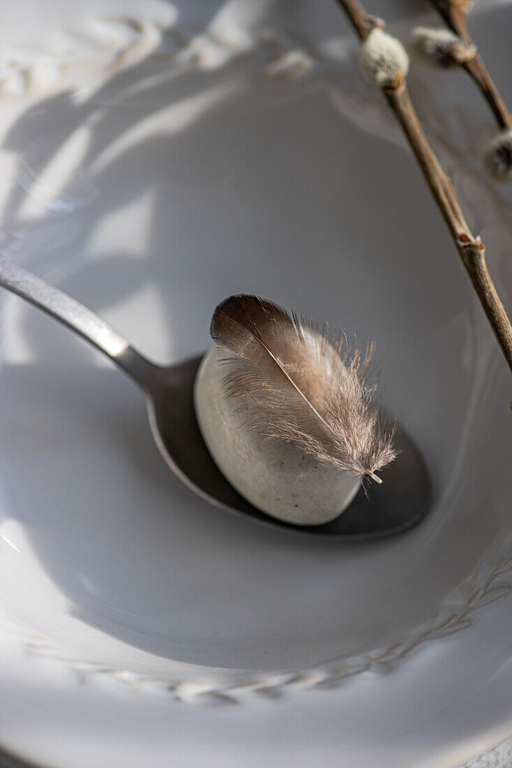 High angle of elegant Easter dining arrangement, featuring pristine white plate with a speckled egg on spoon, accompanied by feather and branches