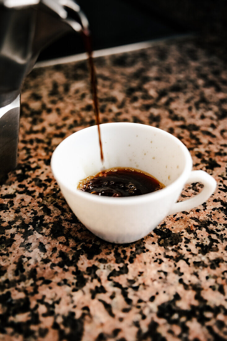 Freshly brewed coffee from a stovetop espresso maker into a white cup set against a modern kitchen backdrop