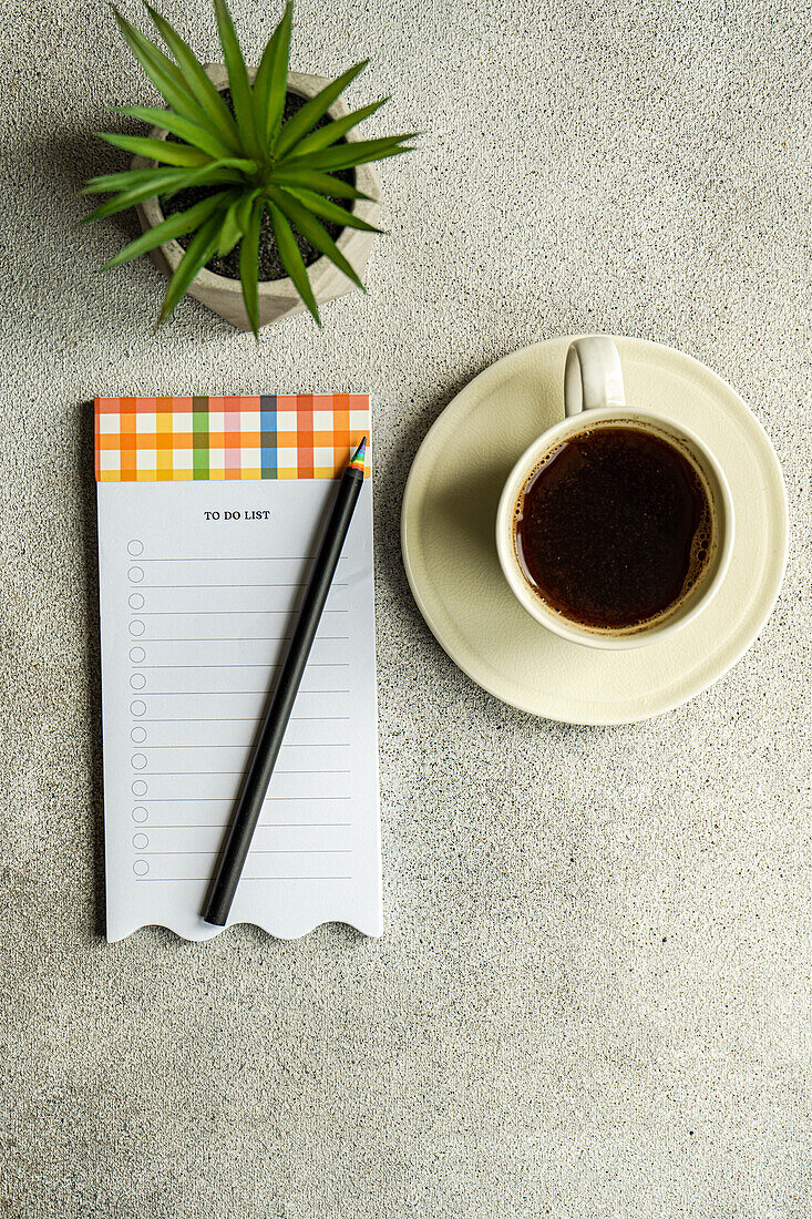 Draufsicht auf einen Kaffeebecher neben einer Topfpflanze und einem Zettel mit Bleistift auf einem grauen Tisch