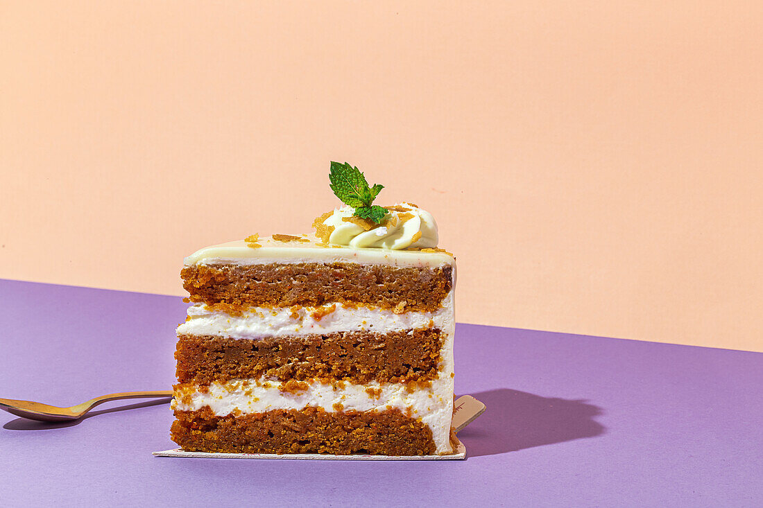 Slice of tasty sweet carrot sponge cake with cream decorated with mint leaf served on plate with spoon on table on colorful background