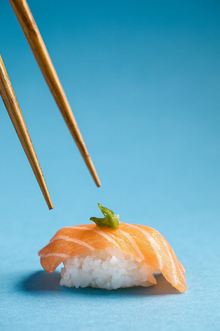 Leckeres japanisches Nigiri mit frischem Lachs und grünem Wasabi vor blauem Hintergrund mit Stäbchen in einem hellen Studio