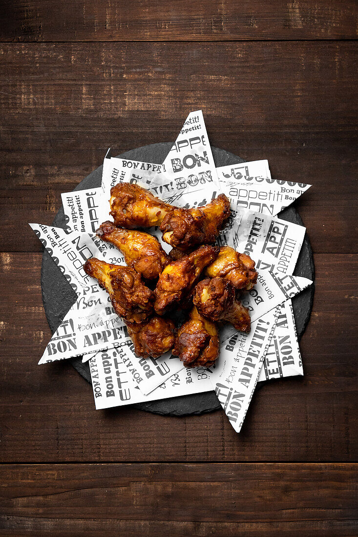 Top view of appetizing roasted chicken wings placed on black plate decorated with newspaper scraps on wooden table