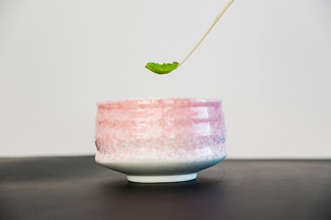 Wooden chashaku of dried green tea adding to ceramic bowl placed on table against white background