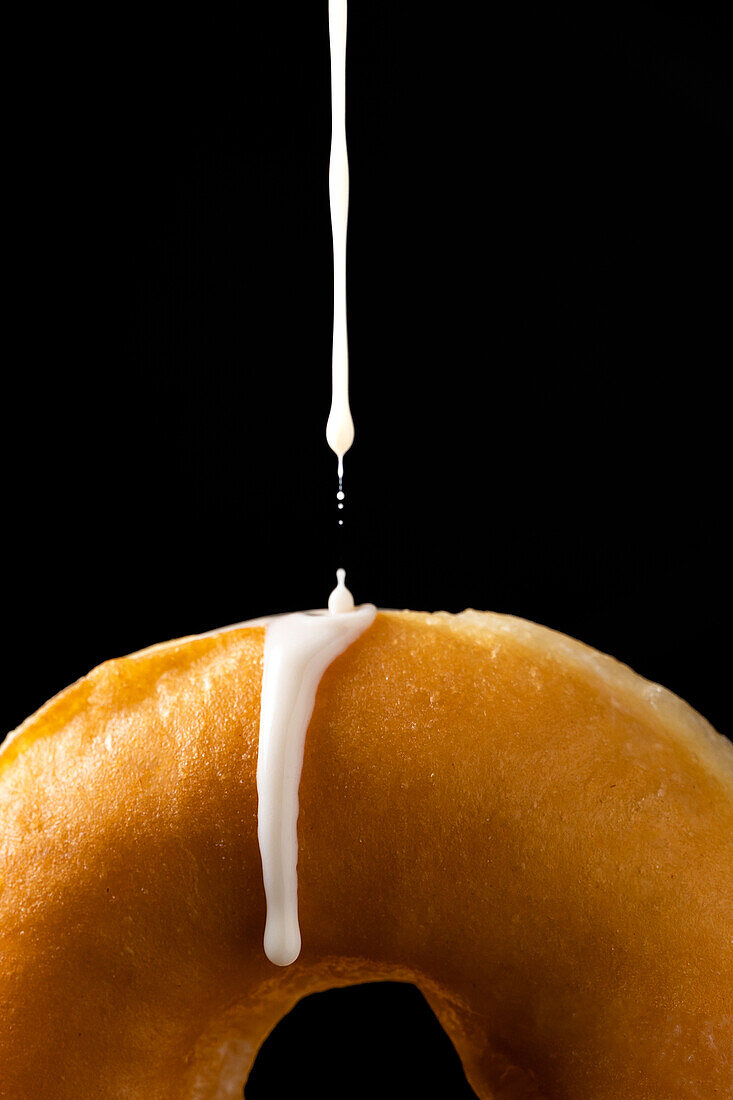 Anonyme Person gießt weißen Zuckerguss Milch auf leckeren süßen Donut vor schwarzem Hintergrund