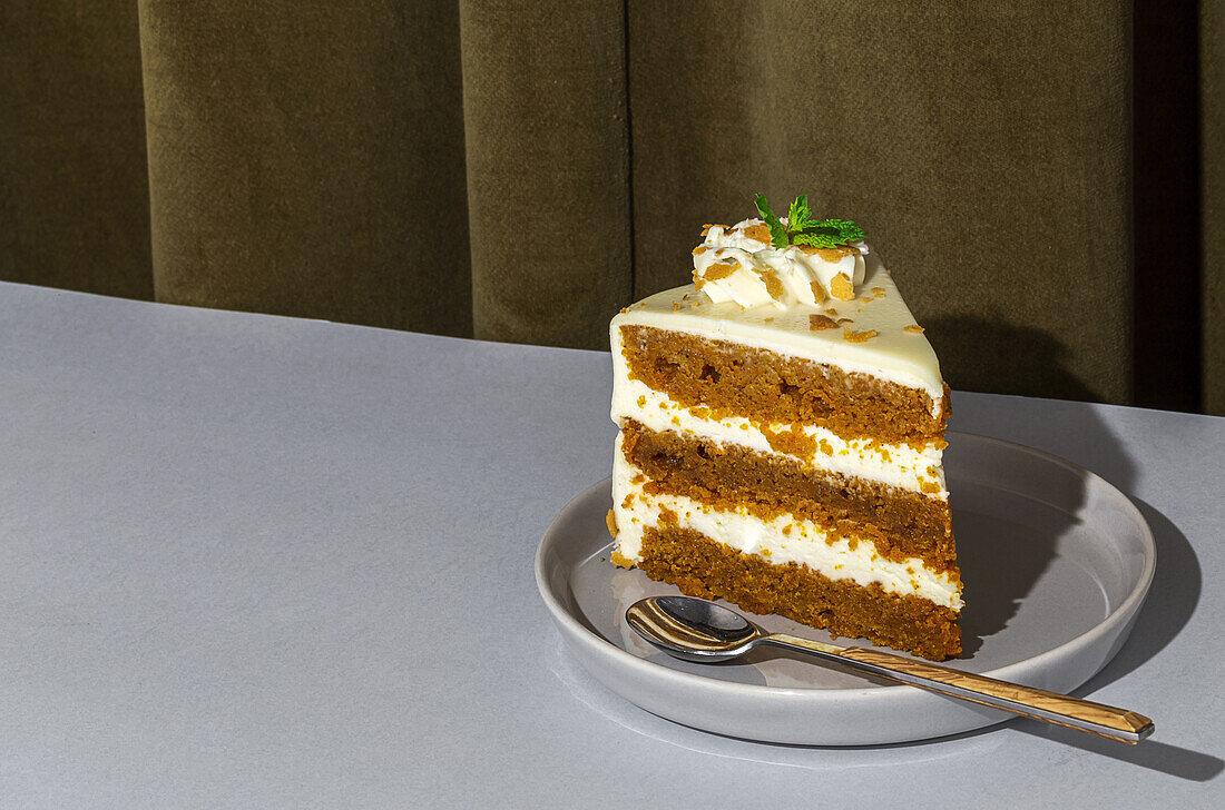 Slice of tasty sweet carrot sponge cake with cream decorated with mint leaf served on plate with spoon on table in light room with curtain on the background