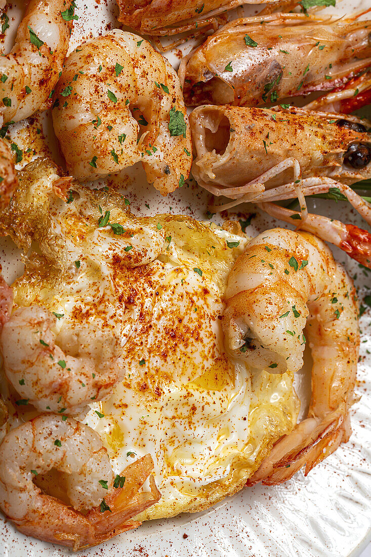 From above appetizing fried eggs with king prawns served on white plate with green herbs and seasoning on towel in light kitchen