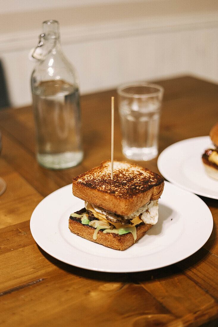 From above of appetizing sandwich with fried toasts placed on white plate and pierced with toothpick while served on wooden table near bottle of water