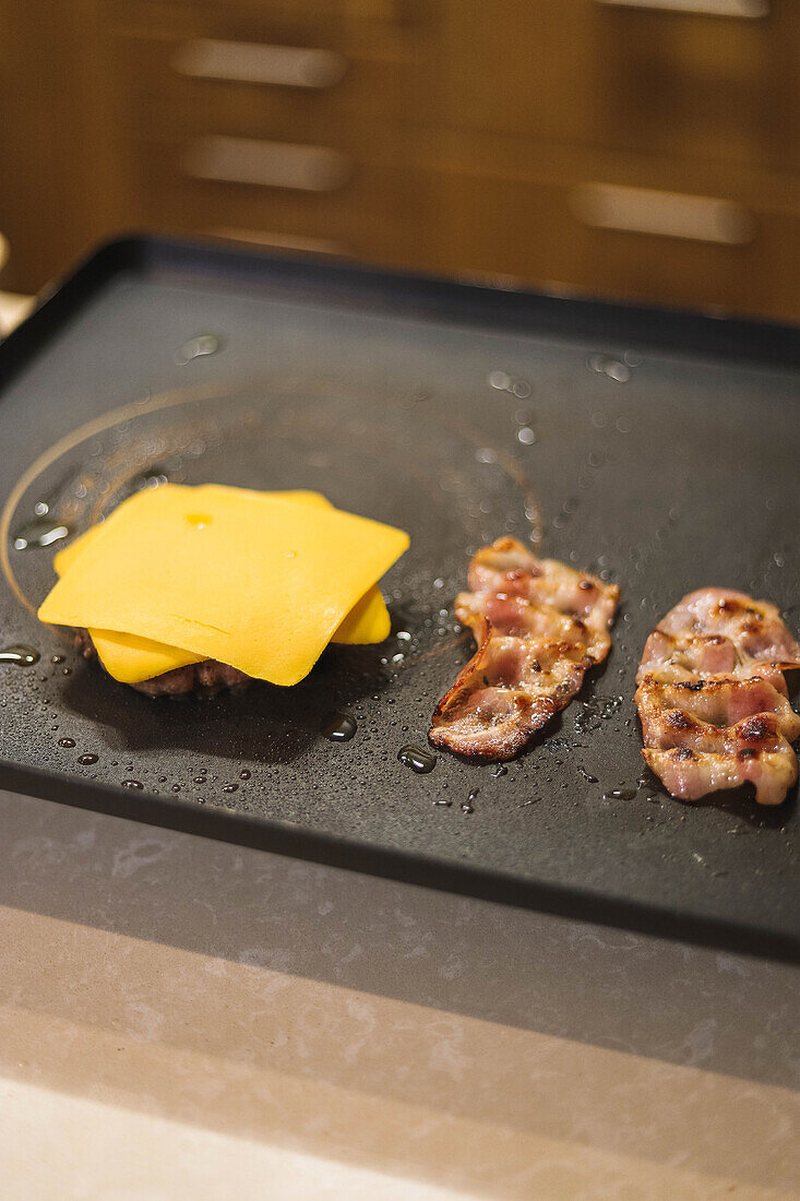 From above grilled patty for burger with cheese on top and bacon slices on black stove