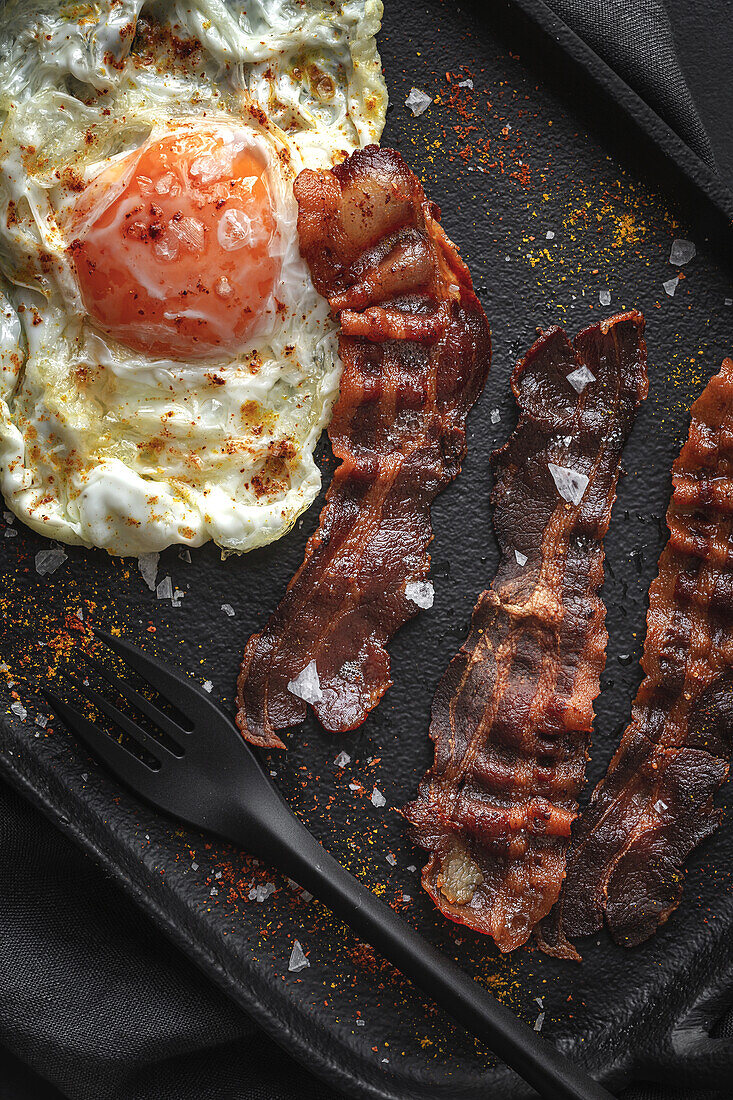 Top view of sunny side up egg with fried bacon slices and condiments on tray against cutlery on dark background