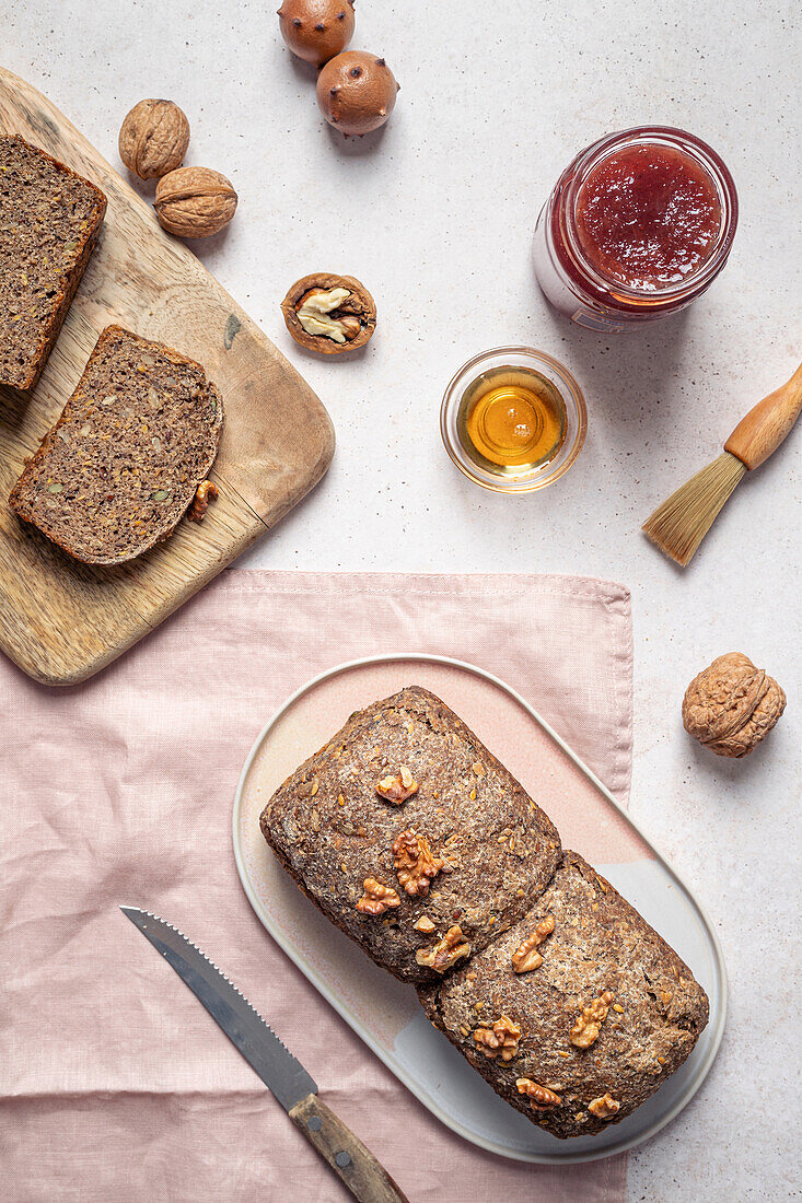 Top view composition of seeded whole grain bread arranged with honey jar of jam and walnut on table near knife