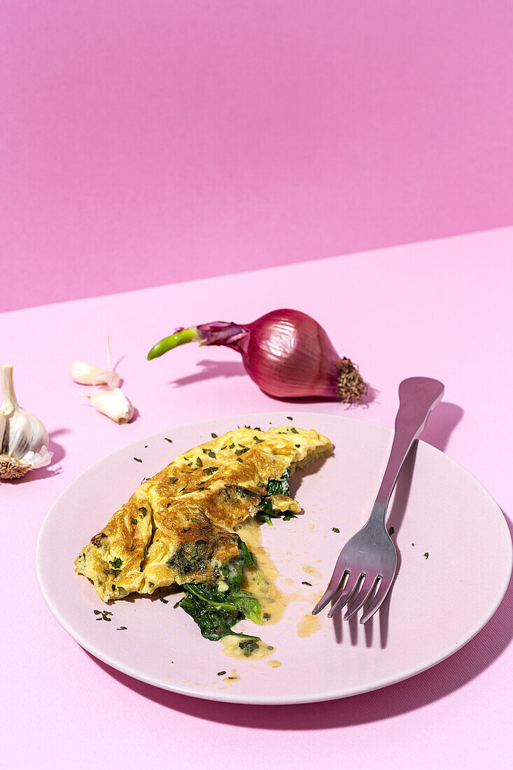 Leckeres Omelett auf einem Teller mit frischen Petersilienzweigen und roten Zwiebeln mit Knoblauchzehen auf rosa Hintergrund