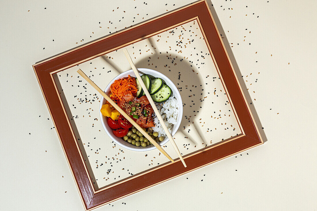 From above white bowl with tasty poke dish and chopsticks placed behind frame on table covered with sesame seeds