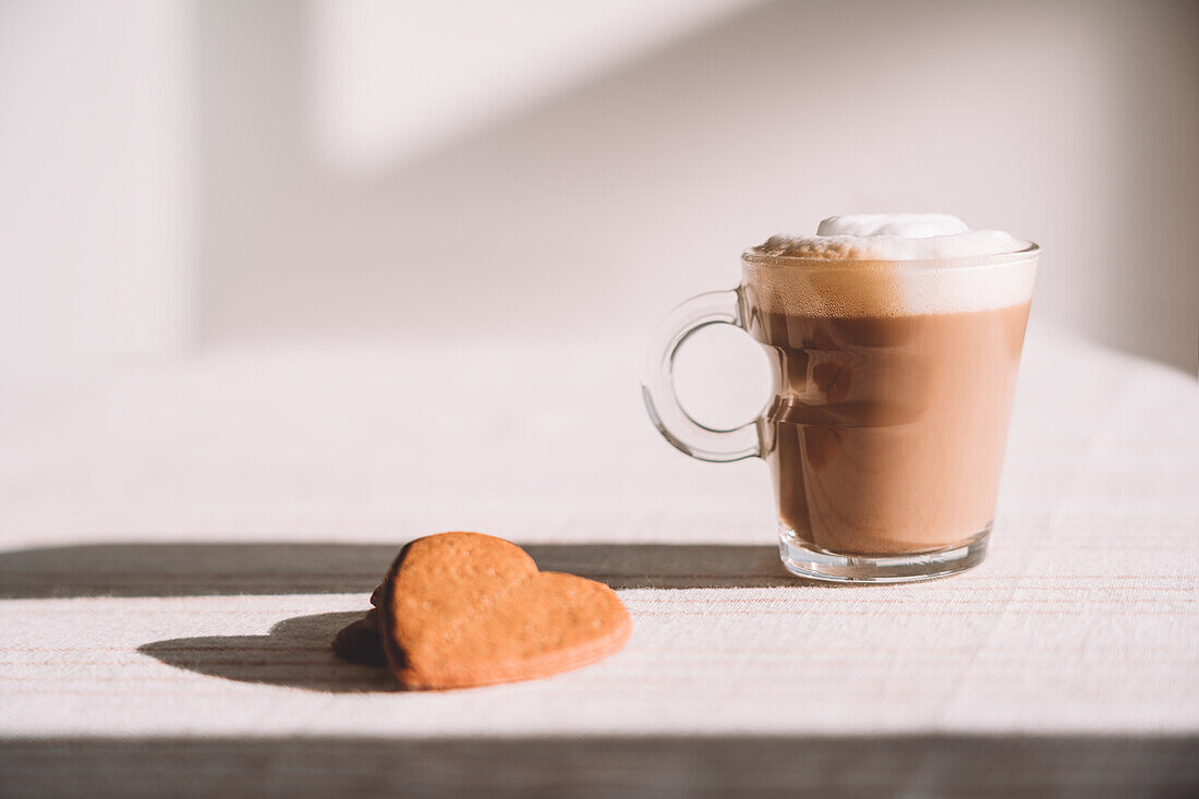 Glass of foamy latte macchiato served on table with heart shaped cookies on sunny day in kitchen