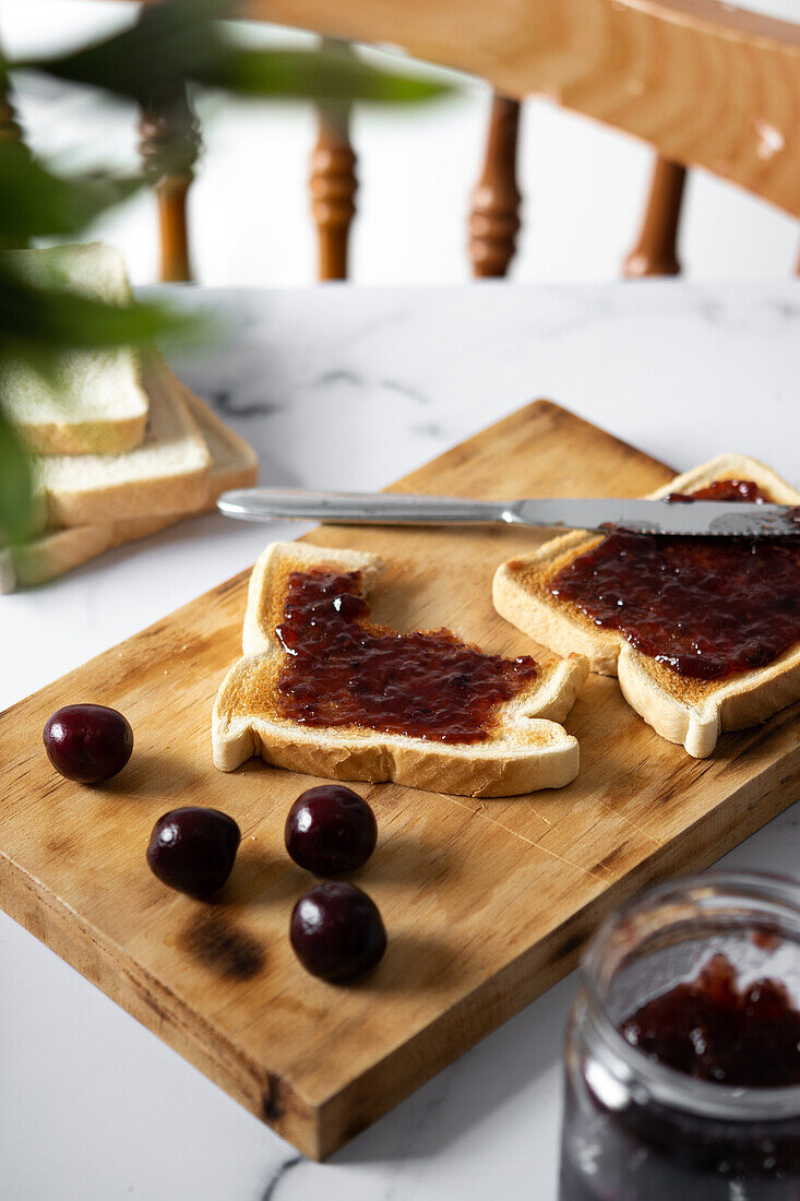 High angle of appetizing crispy toasts with cherry jam served on wooden chopping board near fresh ripe cherries on table