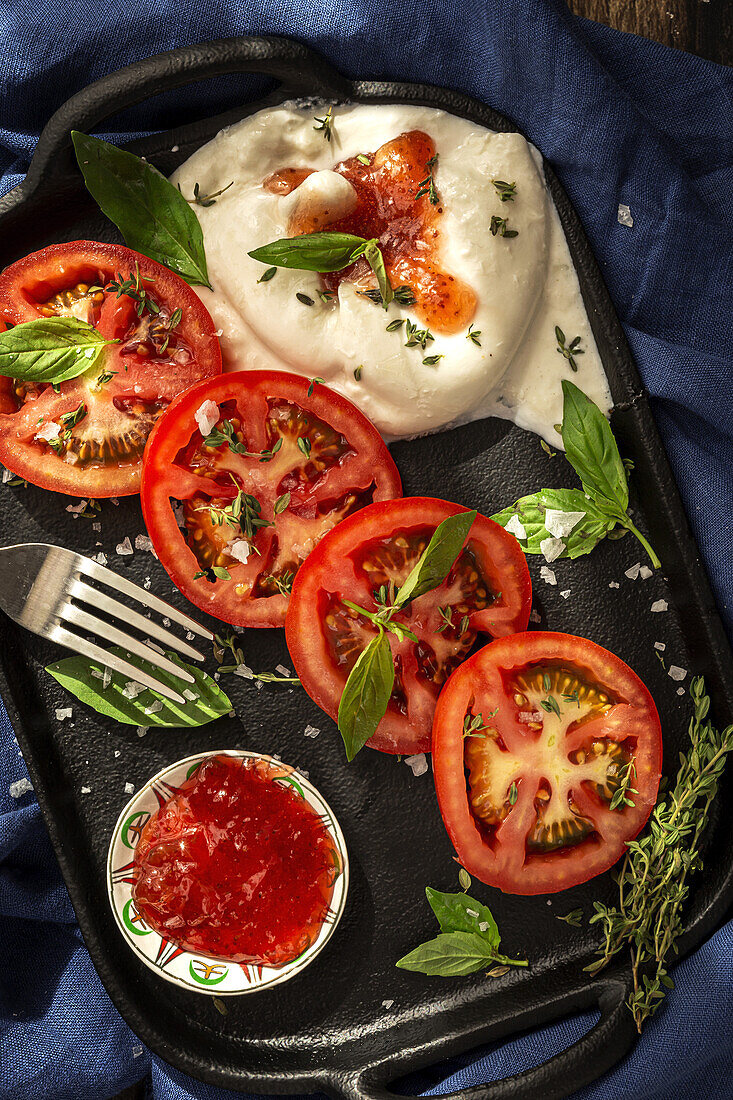 From above of appetizing burrata cheese with sauce and slices of fresh tomato with thyme and basil leaves served on cast iron tray placed on table cloth on wooden table