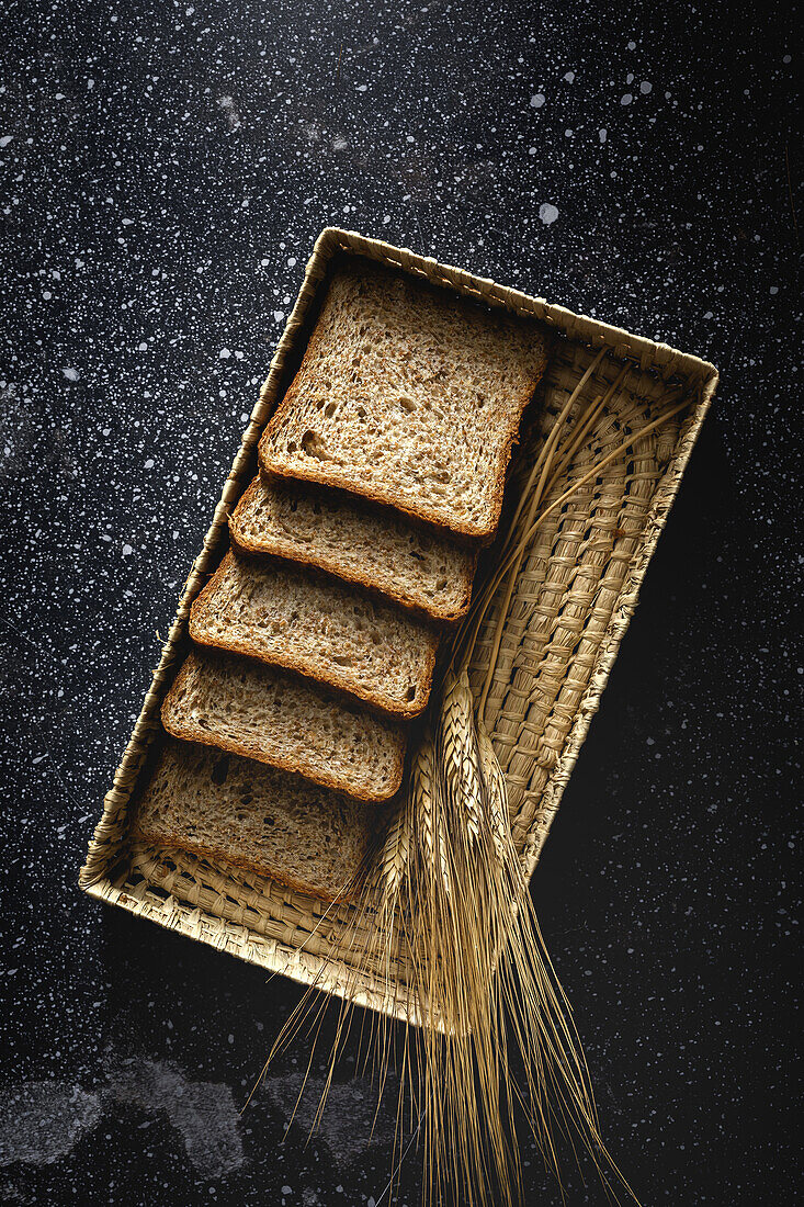 Top view of fresh homemade rye bread near knife in wicker basket and wheat spikes on table