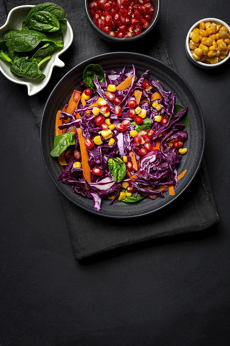 Homemade Purple Cabbage Salad with Corn, Carrots, Pomegranate and Spinach on dark background. Vegan food concept. Healthy food