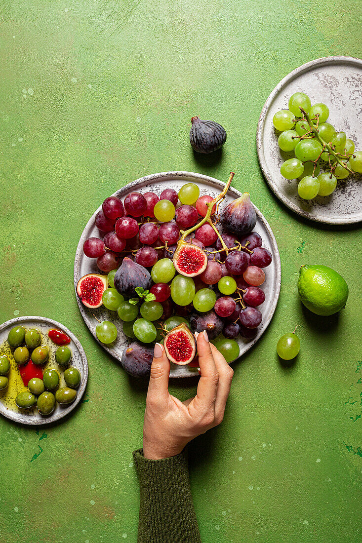 Top view of cropped unrecognizable person eating bunch of delicious fresh juicy red and green grapes served on plate with figs on green background