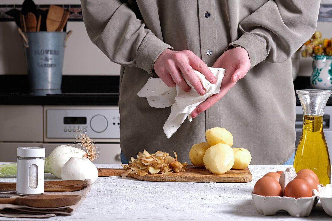 Anonyme Hausfrau wischt sich die Hände mit einer Serviette ab, während sie an einem Tisch mit geschälten Kartoffeln, Zwiebeln und Eiern für ein traditionelles spanisches Omelett-Rezept steht