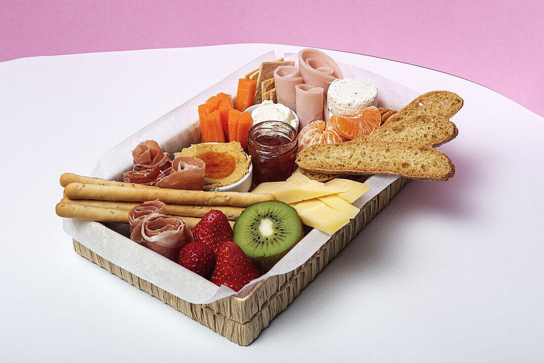 From above brunch box with assorted sliced meats various types of cheese and crispbreads arranged near ripe cup kiwi sweet strawberries and peeled mandarin near jam in glass jar on colorful background