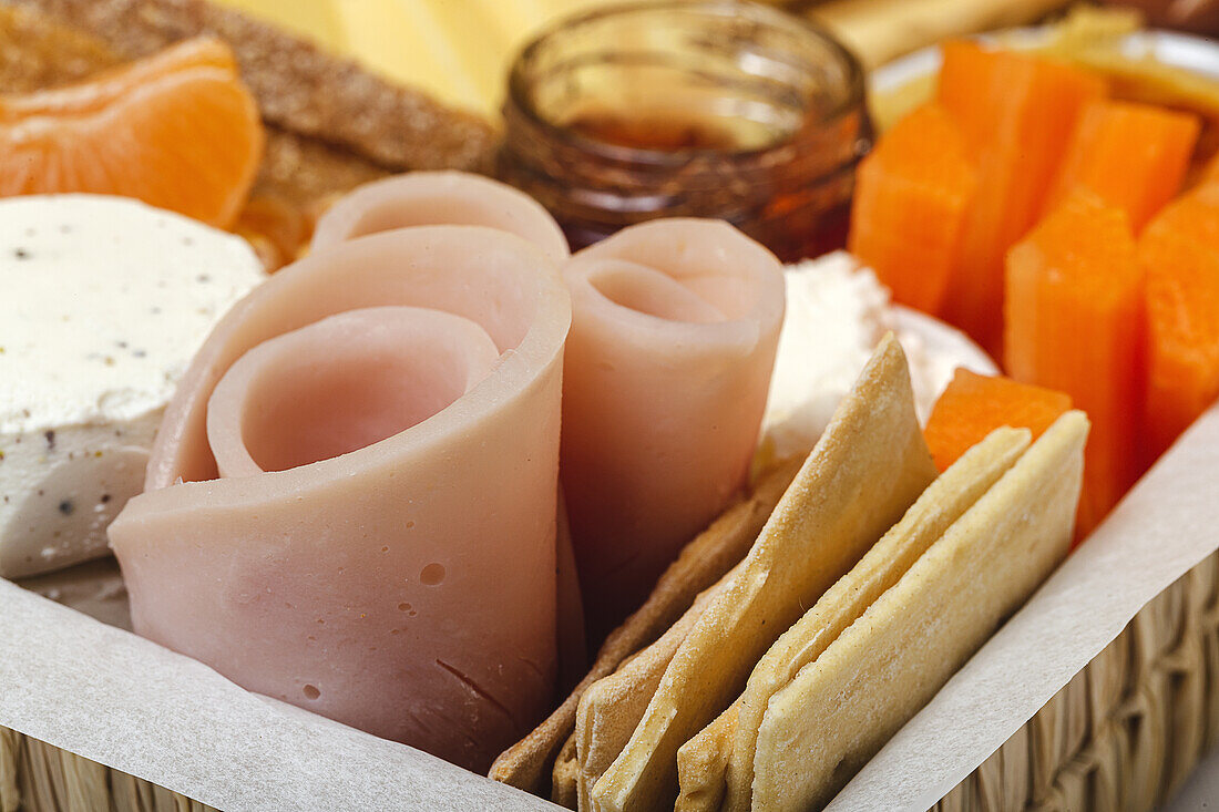 Brunch box with assorted sliced meats various types of cheese and crispbreads arranged near ripe cup kiwi sweet strawberries and peeled mandarin near jam in glass jar on colorful background