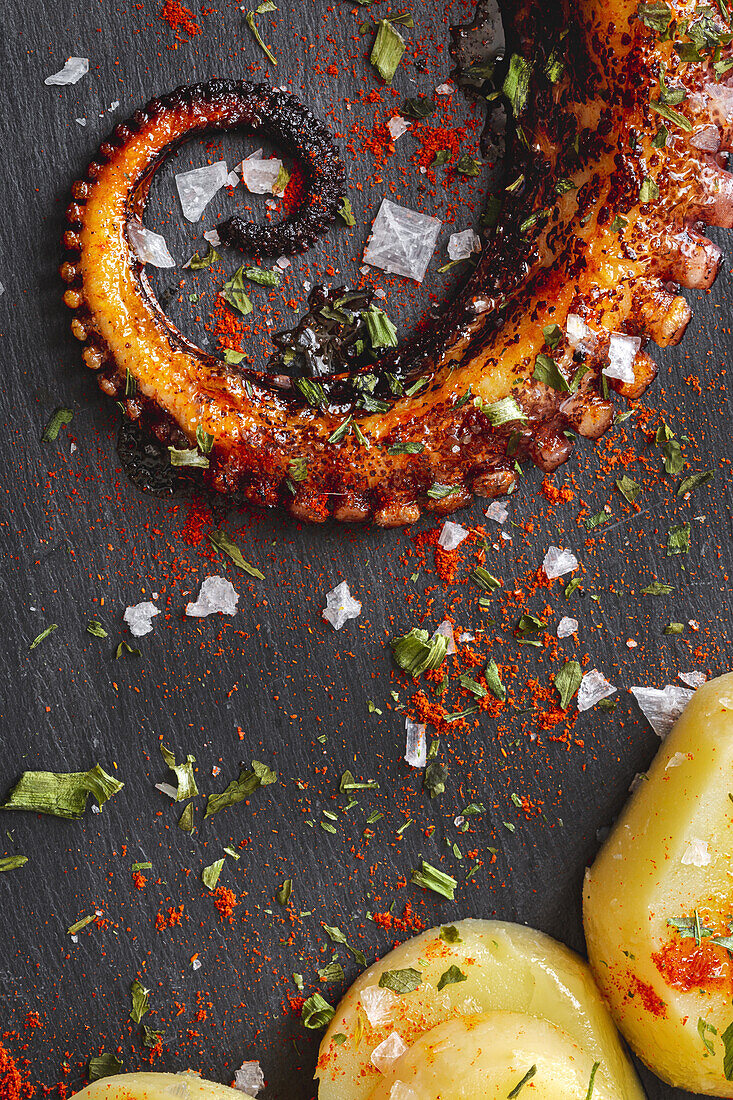 Top view of fried octopus tentacle and pieces of potato served with spices on black board on table