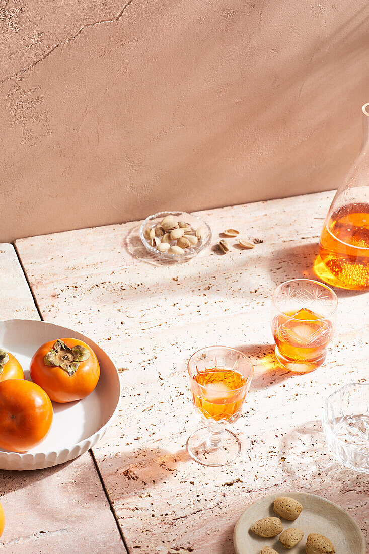 High angle of alcohol cocktail in shot glasses with ice cubes placed on marble tile near persimmon fruits and nuts in sunlight