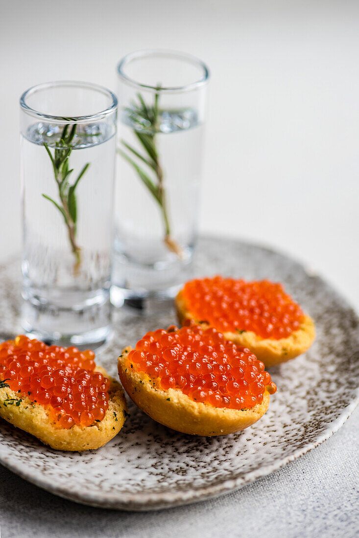 Bruschetta with red caviar in ceramic plate near glasses with rosemary vodka in