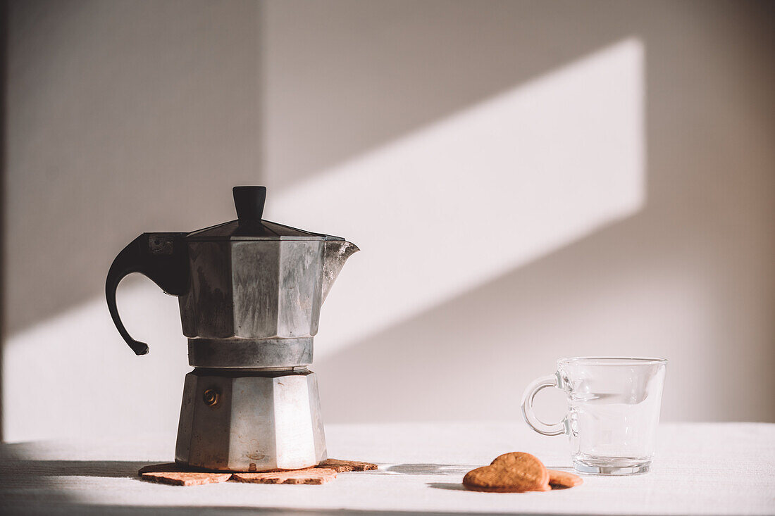 Metall-Moka-Kanne auf weißem Tisch neben Glastasse und herzförmigen Keksen an einem sonnigen Tag