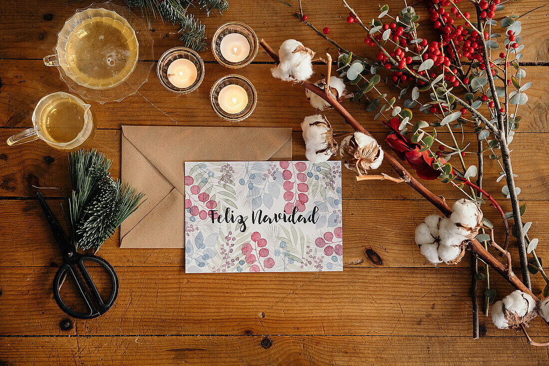 Draufsicht auf eine weihnachtliche Komposition mit einer bunten Postkarte mit der Aufschrift Feliz Navidad in der Nähe von brennenden Kerzen und Teetassen auf einem Holztisch, der mit bunten Pflanzenzweigen dekoriert ist