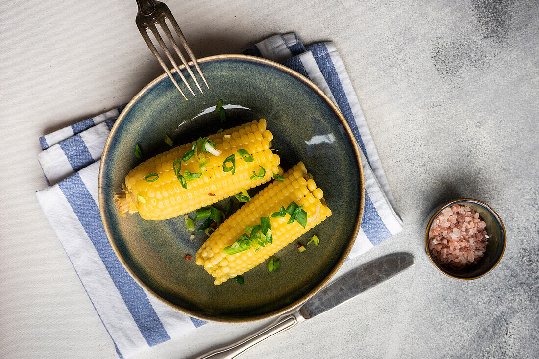 Top view of boiled corn with salt and green onion served on plate on grey background