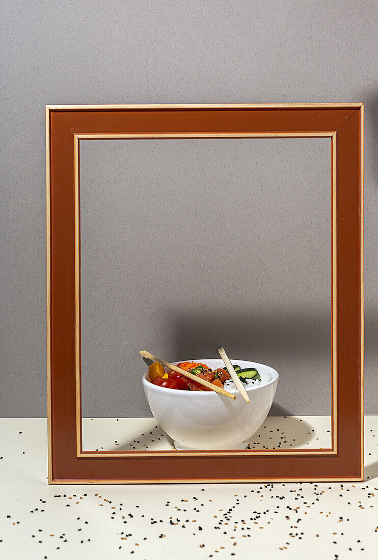Hoher Winkel einer weißen Schale mit leckerem Poke-Gericht und Stäbchen, die hinter dem Rahmen auf einem mit Sesam bedeckten Tisch stehen