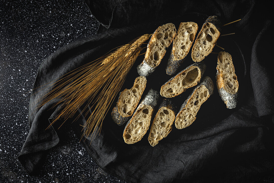 From above of appetizing crusty bread near wheat spikes and dark fabric on table