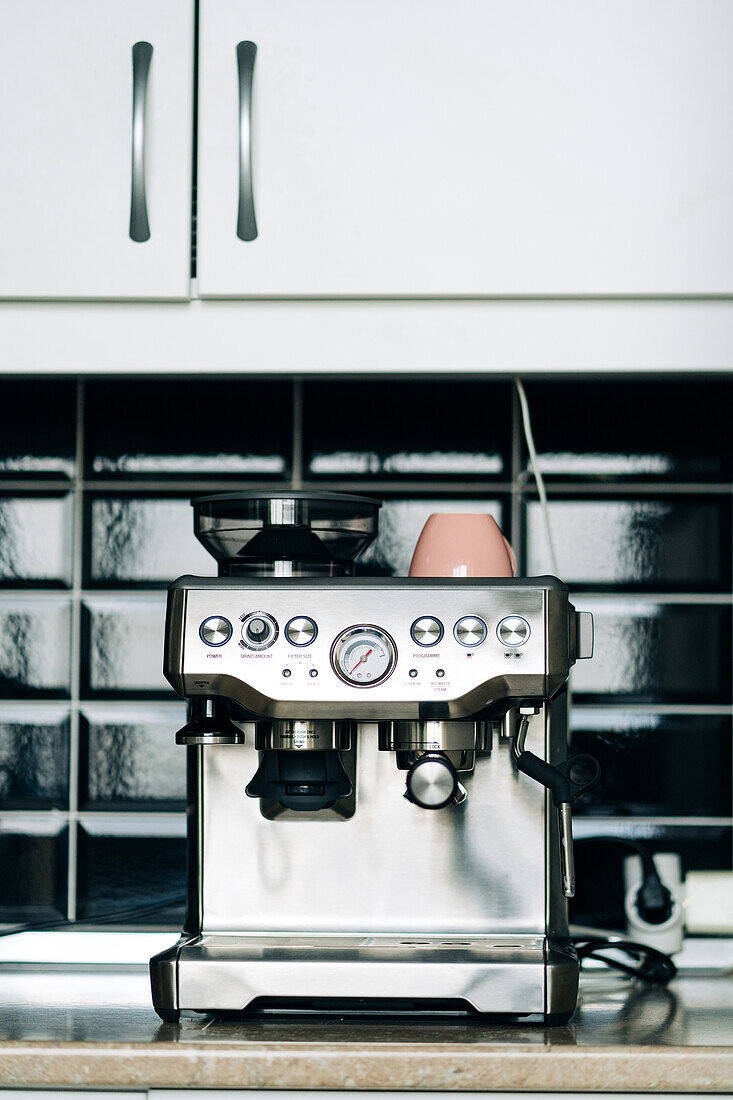 Professionelle Kaffeemaschine aus Edelstahl mit Manometer und Keramiktasse auf einem Tisch vor einer Glasblockwand in einem Haus