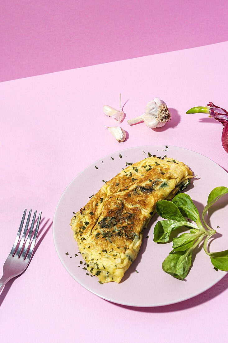 Tasty omelette on plate against fresh parsley sprigs and red onion with garlic cloves on pink background