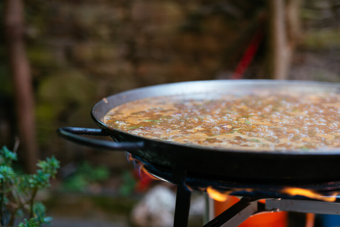 Große Eisenpfanne mit dampfenden Zutaten auf dem Feuer bei der Zubereitung einer traditionellen spanischen Paella im Garten