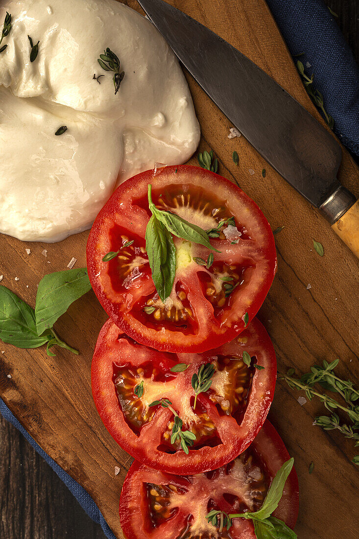 Draufsicht auf köstlichen Mozzarella und Scheiben frischer Tomaten mit grünen Basilikumblättern und Thymian, serviert auf einem hölzernen Schneidebrett mit Messer auf dem Tisch