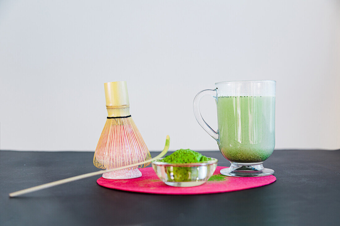 Ein Glas gesunder Matcha-Tee mit Milch in einem Glasbecher und getrocknetem Pulver in der Nähe von hölzernen Chasen und Chashaku auf einem hellen runden Ständer auf einem schwarzen Tisch im Sonnenlicht