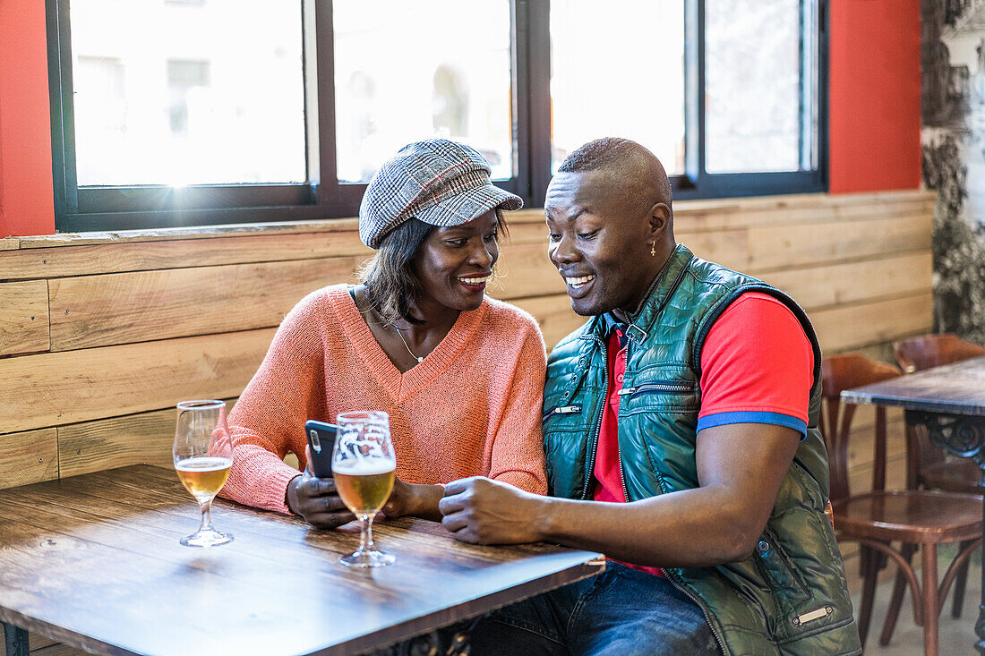 Fröhliches schwarzes Paar benutzt Smartphone, während es am Tisch mit kaltem Bier in einer Bar sitzt