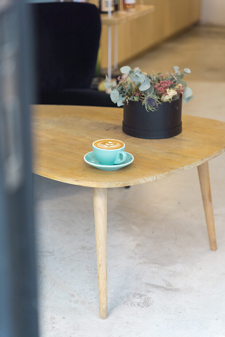 Becher mit heißem Kaffee mit kreativer Latte Art, serviert auf einem Teller mit Teelöffel auf einem Holztisch in einem modernen hellen Kaffeehaus