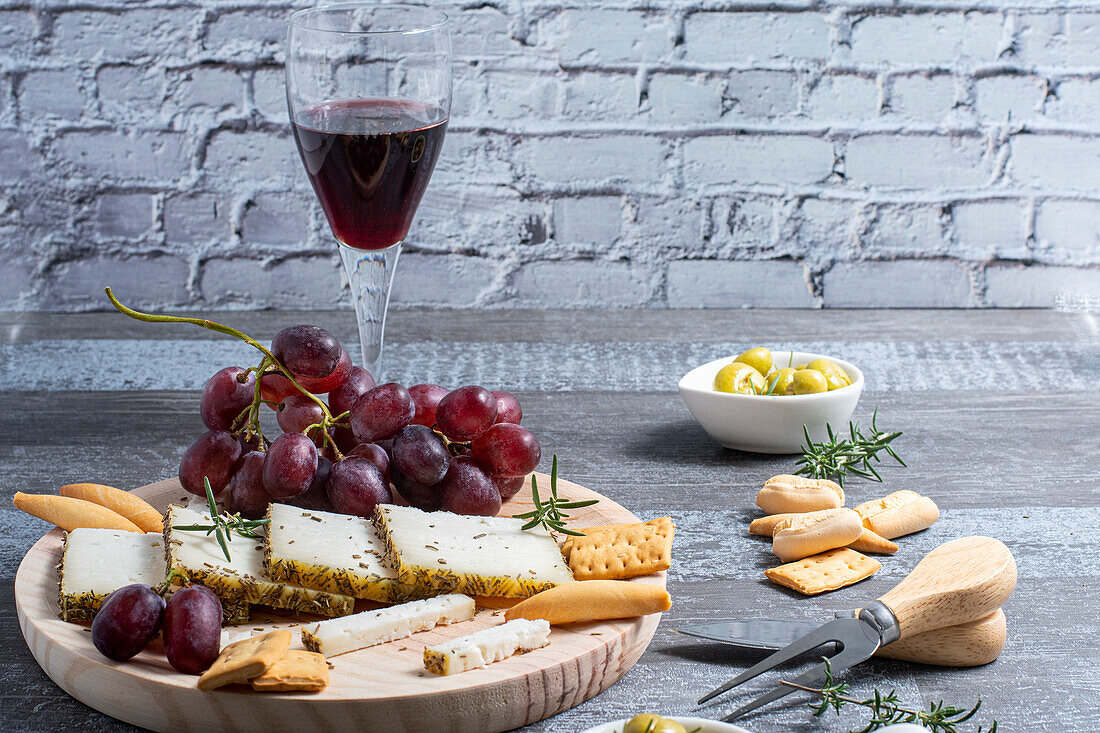 Glass of red wine placed near assorted cheese and grape with crackers and olives decorated with rosemary on table