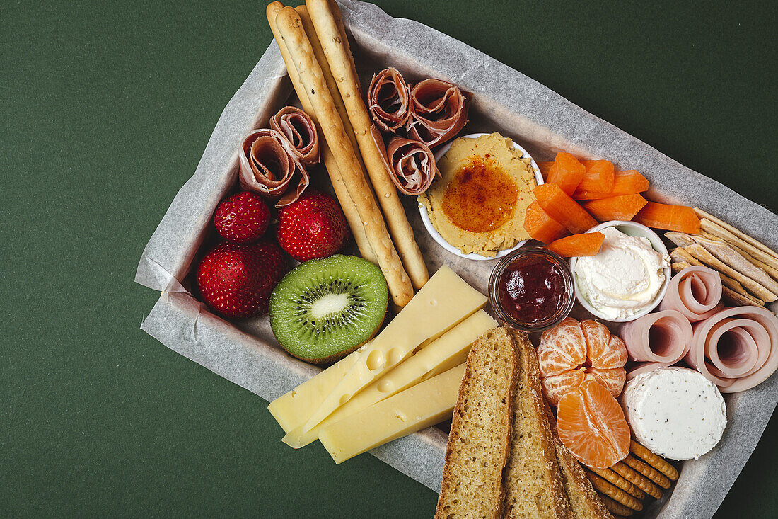 From above brunch box with assorted sliced meats various types of cheese and crispbreads arranged near ripe cup kiwi sweet strawberries and peeled mandarin near jam in glass jar on colorful green background