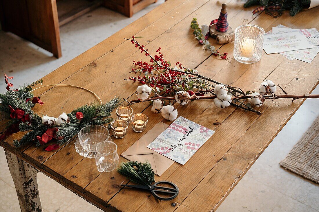 From above Christmas composition with colorful postcard with inscription Feliz Navidad placed near burning candles and cups of tea on wooden table decorated with colorful branches of plants