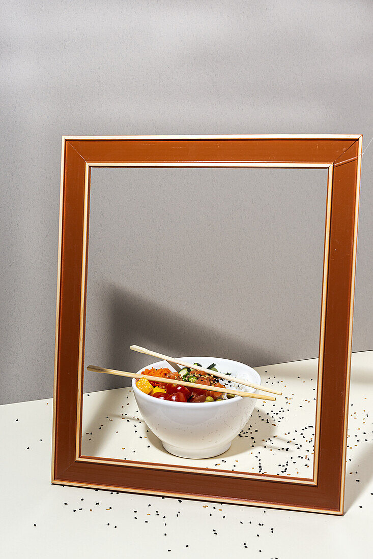 High angle of white bowl with tasty poke dish and chopsticks placed behind frame on table covered with sesame seeds