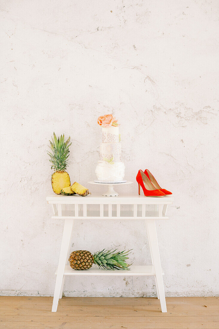 Fresh pineapples and tasty cake placed on table near red high heeled shoes against white wall during wedding celebration