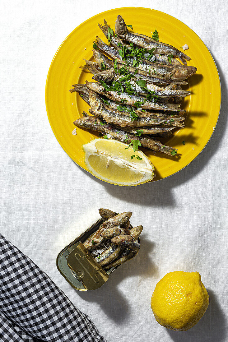 From above of fried and canned anchovies served on table with fresh lemons in restaurant in sunlight