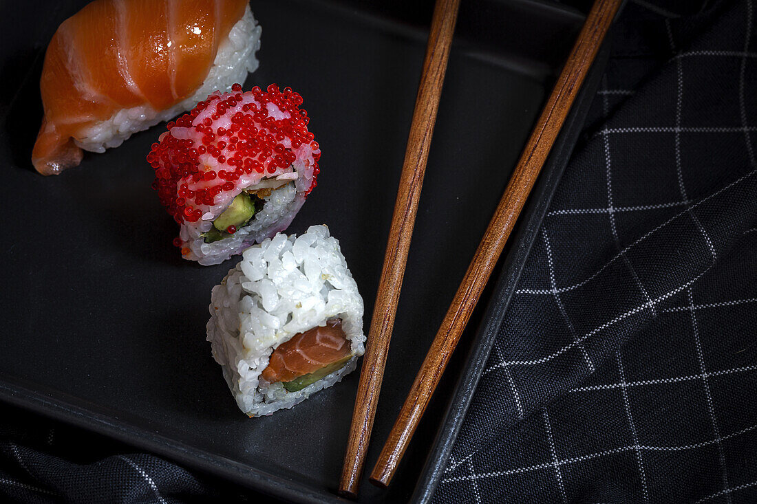 Top view composition of delicious fresh various sushi and bamboo chopsticks served on black platter on checkered cloth